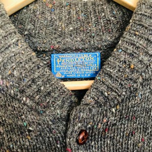 Puede incluir: Primer plano de un c&aacute;rdigan de lana Pendleton gris con cierre de botones. El su&eacute;ter tiene un patr&oacute;n moteado con motas de color. Una etiqueta azul dice "PENDLETON" y "100% VIRGIN WOOL". El su&eacute;ter es talla 2X y est&aacute; hecho en los EE. UU.