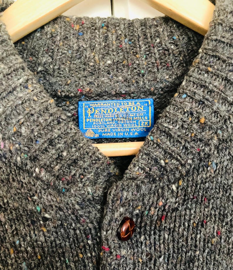 Puede incluir: Primer plano de un c&aacute;rdigan de lana Pendleton gris con cierre de botones. El su&eacute;ter tiene un patr&oacute;n moteado con motas de color. Una etiqueta azul dice "PENDLETON" y "100% VIRGIN WOOL". El su&eacute;ter es talla 2X y est&aacute; hecho en los EE. UU.