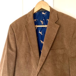 Pu&ograve; includere: Blazer di velluto a coste marrone appeso a una gruccia di legno. Il blazer ha un classico revers a lancia e una tasca frontale. La fodera interna &egrave; blu scuro con un motivo ripetuto di cani marroni e bianchi.