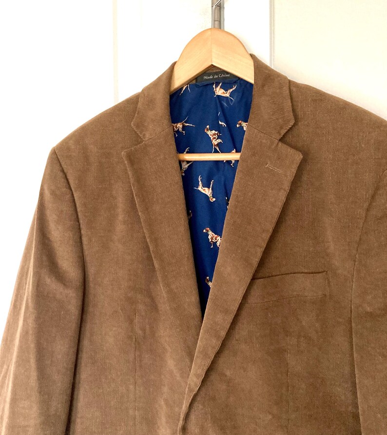 Pu&ograve; includere: Blazer di velluto a coste marrone appeso a una gruccia di legno. Il blazer ha un classico revers a lancia e una tasca frontale. La fodera interna &egrave; blu scuro con un motivo ripetuto di cani marroni e bianchi.