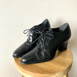 May include: A pair of black leather oxford-style heeled shoes with a lace-up design. The shoes are placed on a small wooden stool. The heels are block style.