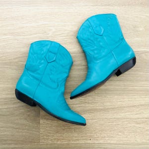 May include: A pair of turquoise cowboy boots. The boots have a pointed toe, a low heel, and decorative stitching. The boots are made of a smooth, shiny material and are set against a light wood grain background.