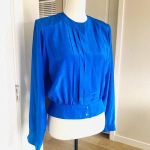 May include: A vibrant blue blouse with long sleeves and a round neckline. The blouse features pleats down the front and buttons at the neckline and waist. The garment is displayed on a mannequin.