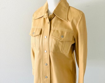 May include: A tan leather jacket with a button-up front and two flap pockets. The jacket has a collar and long sleeves.