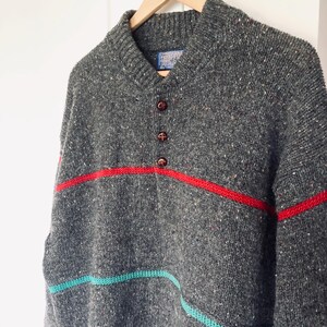 Puede incluir: Un su&eacute;ter de lana gris con un patr&oacute;n moteado, rayas rojas, turquesas y azules, y tres botones marrones. El su&eacute;ter est&aacute; colgado de una percha de madera sobre un fondo blanco. El su&eacute;ter tiene cuello chal.