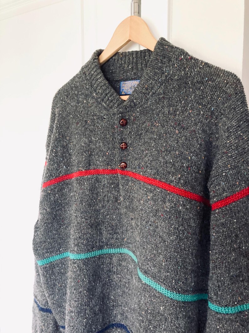 Puede incluir: Un su&eacute;ter de lana gris con un patr&oacute;n moteado, rayas rojas, turquesas y azules, y tres botones marrones. El su&eacute;ter est&aacute; colgado de una percha de madera sobre un fondo blanco. El su&eacute;ter tiene cuello chal.