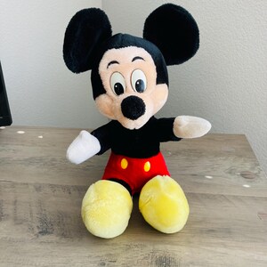 Puede incluir: Un peluche de Mickey Mouse con orejas negras, cabeza negra y cara color canela. El juguete lleva una camisa negra, pantalones cortos rojos con botones amarillos y zapatos amarillos. El juguete est&aacute; sobre una superficie de madera.