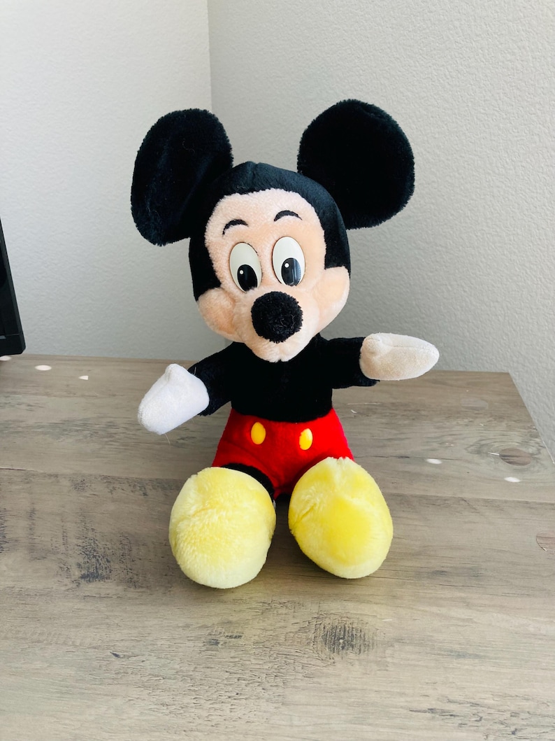 Puede incluir: Un peluche de Mickey Mouse con orejas negras, cabeza negra y cara color canela. El juguete lleva una camisa negra, pantalones cortos rojos con botones amarillos y zapatos amarillos. El juguete est&aacute; sobre una superficie de madera.