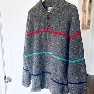 Puede incluir: Un su&eacute;ter gris jaspeado con cuello en V y tres botones. Tiene rayas horizontales en rojo, turquesa y azul. El su&eacute;ter est&aacute; colgado en una percha de madera. Las medidas son en cent&iacute;metros.