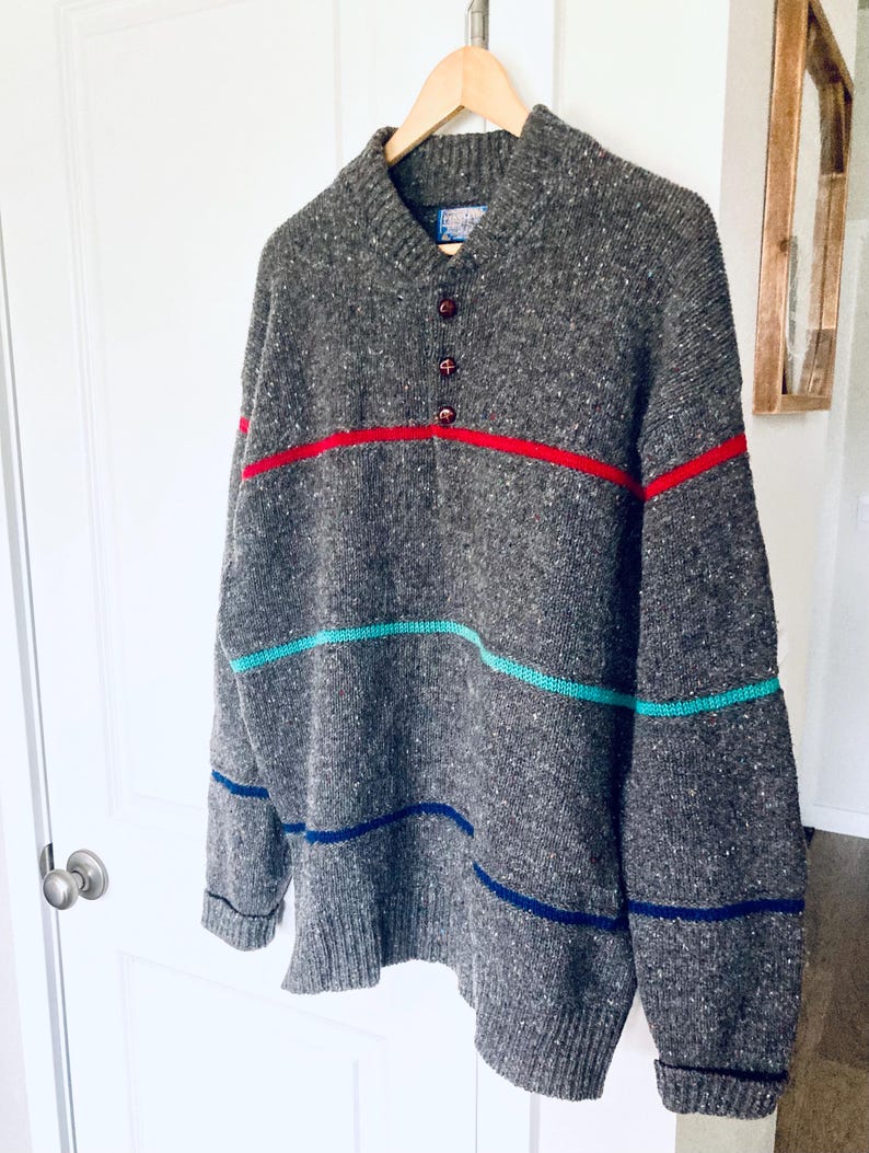 Puede incluir: Un su&eacute;ter gris jaspeado con cuello en V y tres botones. Tiene rayas horizontales en rojo, turquesa y azul. El su&eacute;ter est&aacute; colgado en una percha de madera. Las medidas son en cent&iacute;metros.