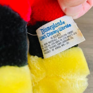Puede incluir: Primer plano de un juguete de peluche con colores rojo, negro y amarillo. El juguete tiene una etiqueta que dice "Disneyland Walt Disney World" y "Disney". La etiqueta tambi&eacute;n incluye informaci&oacute;n sobre los materiales y el origen del producto.
