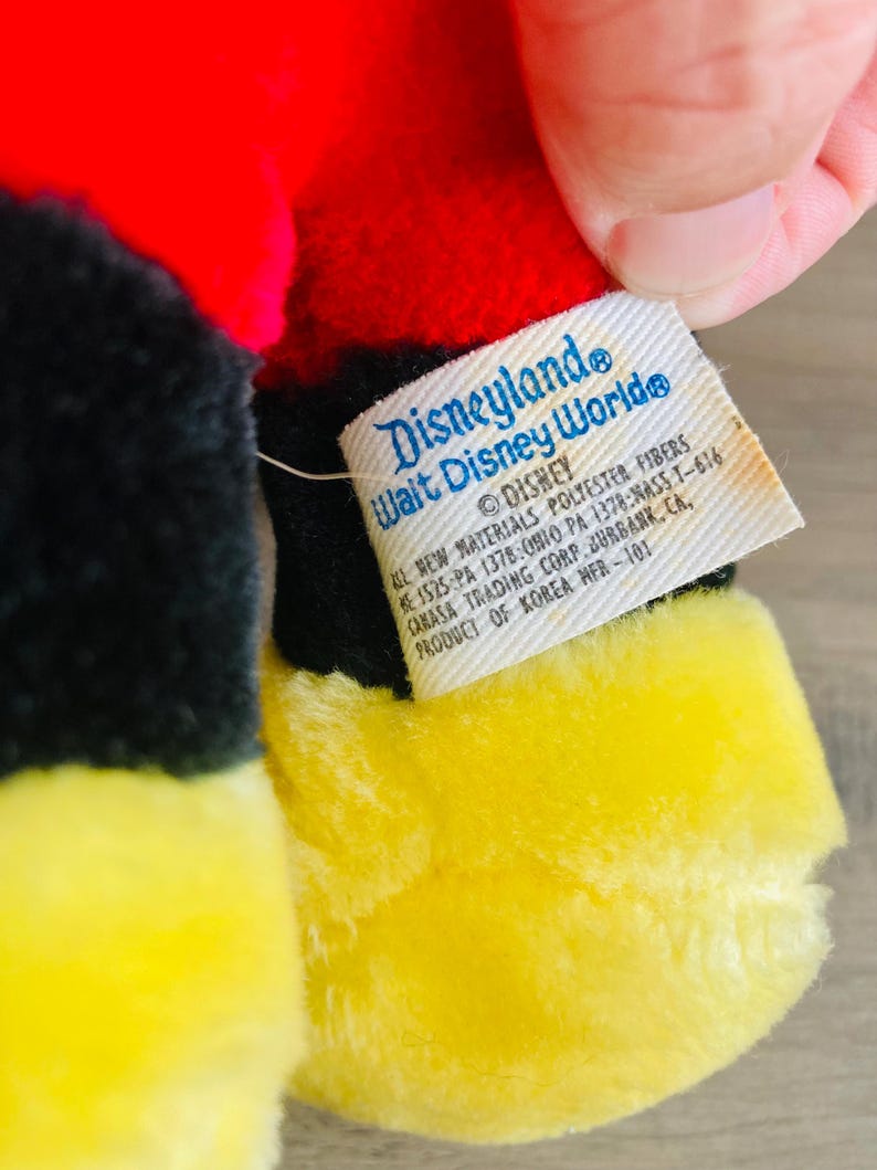 Puede incluir: Primer plano de un juguete de peluche con colores rojo, negro y amarillo. El juguete tiene una etiqueta que dice "Disneyland Walt Disney World" y "Disney". La etiqueta tambi&eacute;n incluye informaci&oacute;n sobre los materiales y el origen del producto.