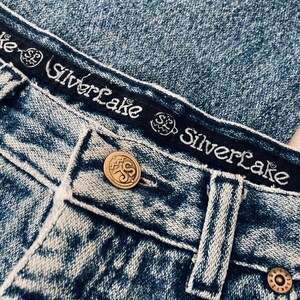 Op de afbeelding: Close-up van een paar blauwe denim jeans met een zwarte tailleband waarop "Silverlake" staat en een gouden knoop met hetzelfde logo. De jeans heeft een verbleekte, verweerde look.