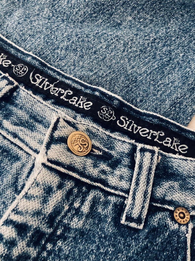 Op de afbeelding: Close-up van een paar blauwe denim jeans met een zwarte tailleband waarop "Silverlake" staat en een gouden knoop met hetzelfde logo. De jeans heeft een verbleekte, verweerde look.