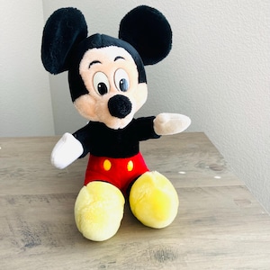 Puede incluir: Un peluche de Mickey Mouse con orejas negras, cabeza negra y cara beige. El juguete tiene un cuerpo negro, pantalones cortos rojos con botones amarillos y pies amarillos. El juguete est&aacute; sobre una superficie de madera.