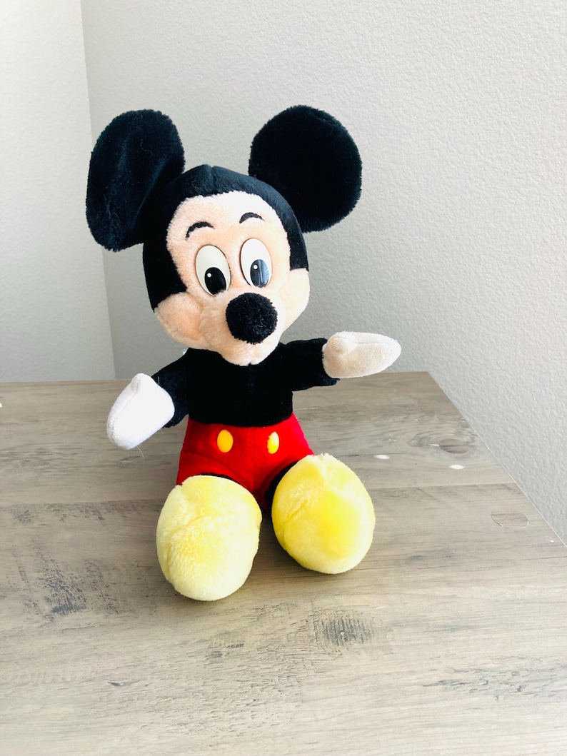 Puede incluir: Un peluche de Mickey Mouse con orejas negras, cabeza negra y cara beige. El juguete tiene un cuerpo negro, pantalones cortos rojos con botones amarillos y pies amarillos. El juguete est&aacute; sobre una superficie de madera.