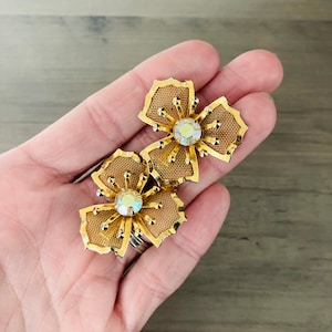 May include: A pair of gold tone flower-shaped earrings with a clear rhinestone in the center of each flower.