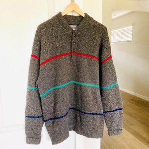 Puede incluir: Un su&eacute;ter de punto gris con cuello abotonado y tres rayas horizontales en rojo, turquesa y azul marino. El su&eacute;ter est&aacute; colgado en una percha de madera contra una puerta blanca.