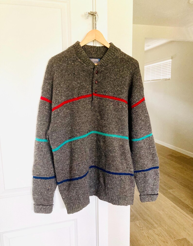 Puede incluir: Un su&eacute;ter de punto gris con cuello abotonado y tres rayas horizontales en rojo, turquesa y azul marino. El su&eacute;ter est&aacute; colgado en una percha de madera contra una puerta blanca.