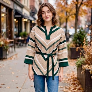 May include: A long-sleeve tunic featuring a chevron pattern in beige, white, and dark green. The garment has a V-neckline, three-quarter length sleeves with dark green cuffs, and a matching dark green fabric tie belt at the waist, paired with blue jeans.