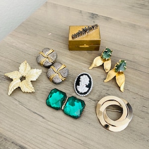 May include: A collection of vintage jewelry pieces, including a gold starburst brooch, gold and silver earrings, a cameo pendant, emerald-cut green gemstone earrings, and a gold and silver brooch. A small gold box is also present.