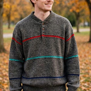 May include: A charcoal gray speckled knit sweater featuring a henley neckline with three brown buttons. The sweater includes horizontal stripes in red, teal, and navy blue across the chest and sleeves, finished with a ribbed collar and cuffs.