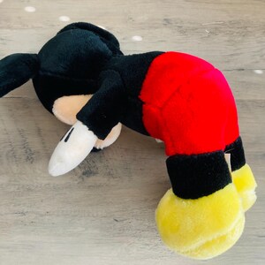 Puede incluir: Un peluche de Mickey Mouse en posici&oacute;n acostada. El juguete presenta una cabeza y cuerpo negros, pantalones cortos rojos, pies amarillos y guantes blancos. Est&aacute; sobre una superficie de madera clara.