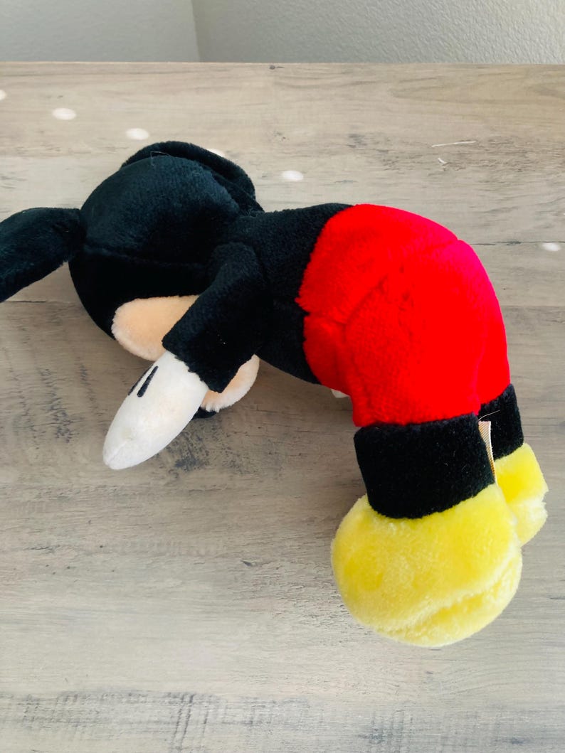 Puede incluir: Un peluche de Mickey Mouse en posici&oacute;n acostada. El juguete presenta una cabeza y cuerpo negros, pantalones cortos rojos, pies amarillos y guantes blancos. Est&aacute; sobre una superficie de madera clara.