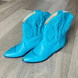 May include: A pair of turquoise cowboy boots. The boots are made of leather and feature decorative stitching on the shaft. The interior is lined with a light-colored material. The boots have a pointed toe and a low heel.