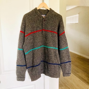 Puede incluir: Un su&eacute;ter marr&oacute;n moteado con cuello abotonado y rayas horizontales en rojo, turquesa y azul marino. El su&eacute;ter est&aacute; colgado de una percha de madera.