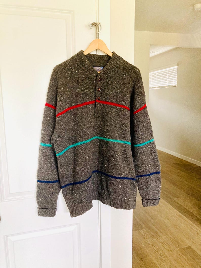 Puede incluir: Un su&eacute;ter marr&oacute;n moteado con cuello abotonado y rayas horizontales en rojo, turquesa y azul marino. El su&eacute;ter est&aacute; colgado de una percha de madera.