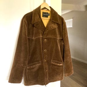 May include: A brown corduroy jacket with a notched collar, three buttons, and two front pockets. The jacket is hanging on a wooden hanger. The interior lining is a light yellow color. The jacket has the brand name "Towncraft" on the label.