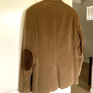 Pu&ograve; includere: Un blazer di velluto a coste marrone con toppe sui gomiti marroni scuri &egrave; appeso a una gruccia di legno. Il blazer ha tre bottoni su ogni polsino e un design classico su misura. La giacca &egrave; esposta contro una porta bianca.