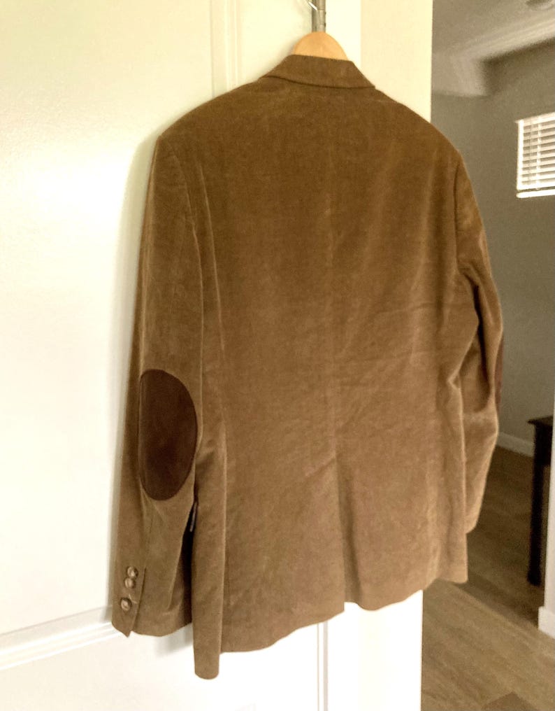 Pu&ograve; includere: Un blazer di velluto a coste marrone con toppe sui gomiti marroni scuri &egrave; appeso a una gruccia di legno. Il blazer ha tre bottoni su ogni polsino e un design classico su misura. La giacca &egrave; esposta contro una porta bianca.