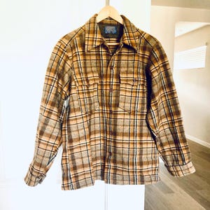 May include: A brown, tan, and black plaid button-up shirt with two chest pockets. The shirt is hanging on a wooden hanger. The shirt has a collar and buttoned cuffs. The shirt is made of a thick, woven fabric.