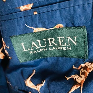 Pu&ograve; includere: Primo piano di un tessuto blu con un motivo ripetuto di cani in varie pose. Il tessuto presenta un'etichetta verde con la scritta "LAUREN RALPH LAUREN" in bianco. I cani sono marroni e bianchi.