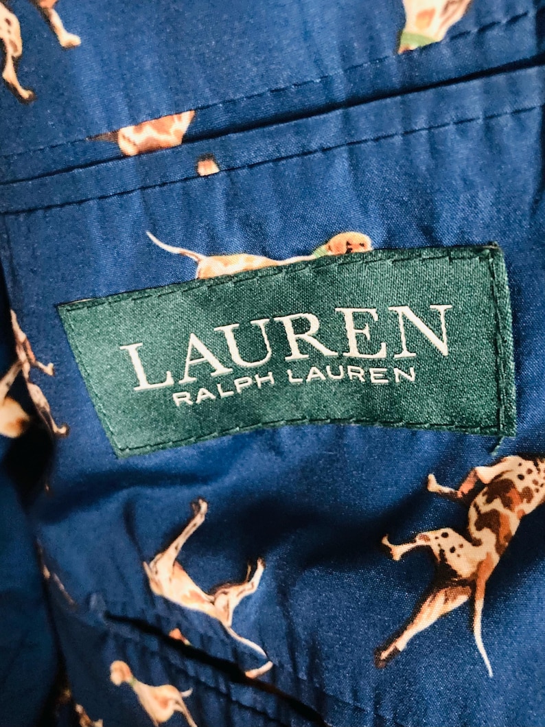 Pu&ograve; includere: Primo piano di un tessuto blu con un motivo ripetuto di cani in varie pose. Il tessuto presenta un'etichetta verde con la scritta "LAUREN RALPH LAUREN" in bianco. I cani sono marroni e bianchi.