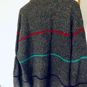 Puede incluir: Un su&eacute;ter gris con rayas horizontales en rojo, turquesa y azul marino. El su&eacute;ter est&aacute; colgado de una percha de madera. El su&eacute;ter tiene una textura moteada.