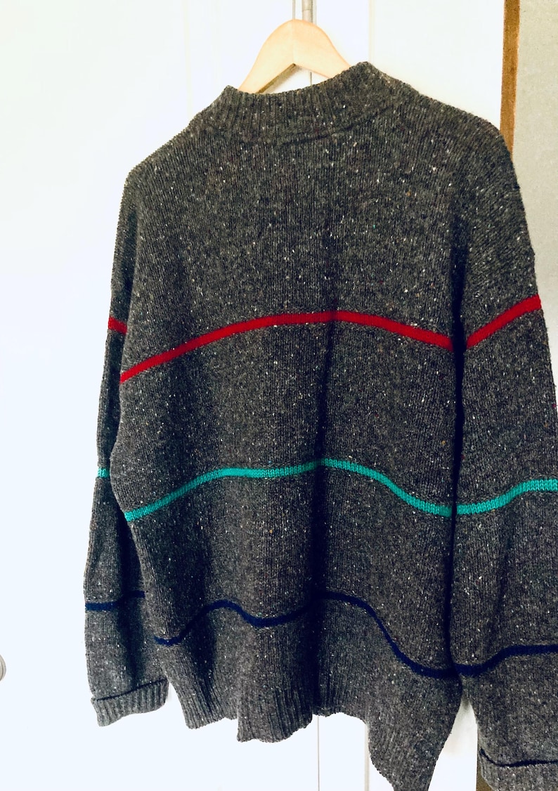 Puede incluir: Un su&eacute;ter gris con rayas horizontales en rojo, turquesa y azul marino. El su&eacute;ter est&aacute; colgado de una percha de madera. El su&eacute;ter tiene una textura moteada.