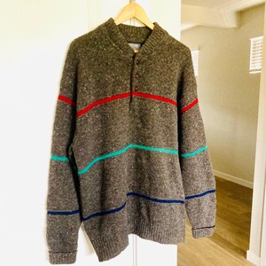 Puede incluir: Un su&eacute;ter gris de manga larga con cuello abotonado y rayas horizontales en rojo, turquesa y azul marino. El su&eacute;ter est&aacute; colgado en una percha de madera contra una puerta blanca.