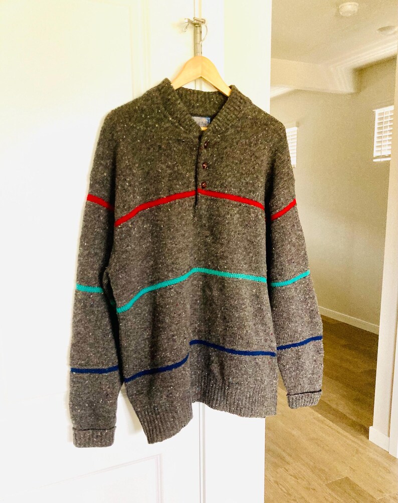 Puede incluir: Un su&eacute;ter gris de manga larga con cuello abotonado y rayas horizontales en rojo, turquesa y azul marino. El su&eacute;ter est&aacute; colgado en una percha de madera contra una puerta blanca.