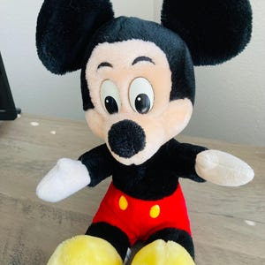 Puede incluir: Un peluche de Mickey Mouse. El juguete tiene cabeza y orejas negras, cara bronceada, guantes blancos, cuerpo negro, pantalones cortos rojos con botones amarillos y pies amarillos. El juguete est&aacute; sobre una superficie de madera.