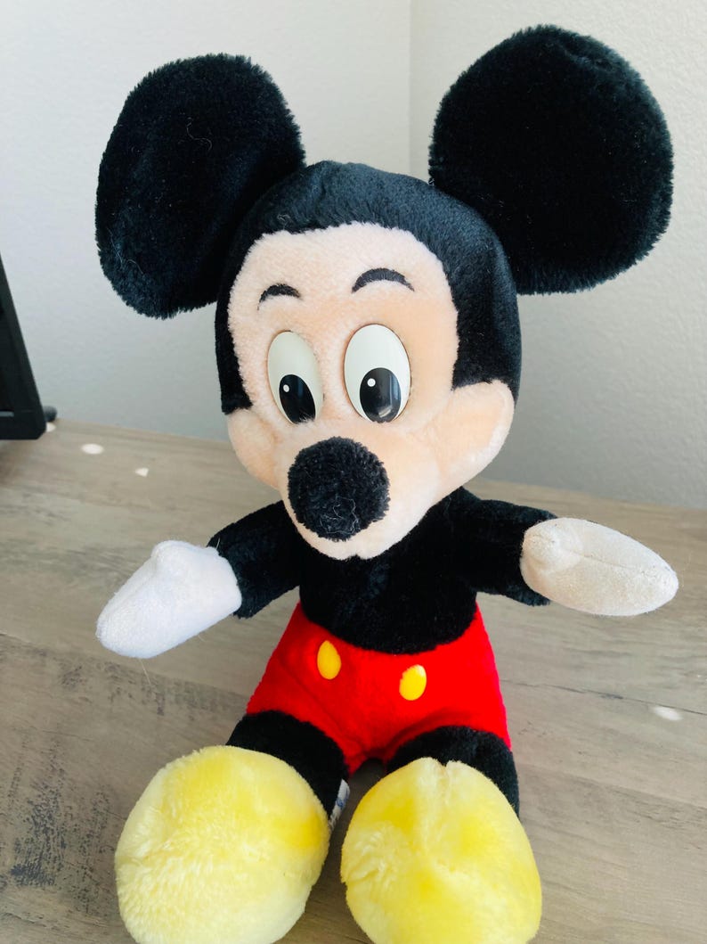 Puede incluir: Un peluche de Mickey Mouse. El juguete tiene cabeza y orejas negras, cara bronceada, guantes blancos, cuerpo negro, pantalones cortos rojos con botones amarillos y pies amarillos. El juguete est&aacute; sobre una superficie de madera.