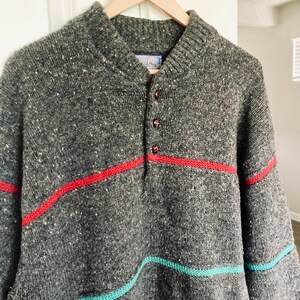 Puede incluir: Un su&eacute;ter de punto gris con rayas rojas y turquesas. El su&eacute;ter tiene un cuello abotonado y est&aacute; colgado de una percha de madera. El su&eacute;ter tiene una textura moteada y un cuello acanalado.