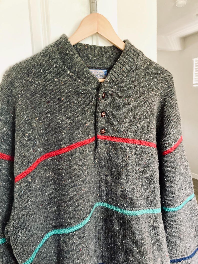 Puede incluir: Un su&eacute;ter de punto gris con rayas rojas y turquesas. El su&eacute;ter tiene un cuello abotonado y est&aacute; colgado de una percha de madera. El su&eacute;ter tiene una textura moteada y un cuello acanalado.