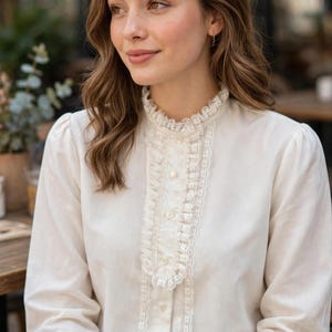 May include: An off-white long-sleeved blouse featuring a high ruffled collar and a vertical lace-trimmed placket down the center front. The garment includes a button-down closure with small round buttons and gathered cuffs at the wrists.