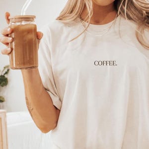 Könnte beinhalten: Eine Person trägt ein cremefarbenes T-Shirt mit dem Aufdruck "COFFEE" auf der Vorderseite und hält ein Glasgefäß mit einem Strohhalm, das mit einem braunen Getränk gefüllt ist. Die Person trägt weiße Socken mit schwarzen Streifen.
