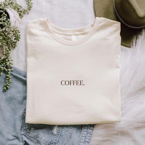 K&ouml;nnte beinhalten: Cremefarbenes T-Shirt mit dem Wort "COFFEE" in schwarzer Schrift. Das Shirt liegt gefaltet auf einer wei&szlig;en, strukturierten Oberfl&auml;che, neben einer Jeans, einem gr&uuml;nen Hut und einer Topfpflanze.