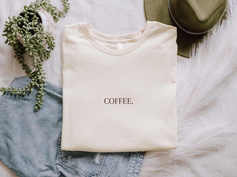 K&ouml;nnte beinhalten: Cremefarbenes T-Shirt mit dem Wort "COFFEE" in schwarzer Schrift. Das Shirt liegt gefaltet auf einer wei&szlig;en, strukturierten Oberfl&auml;che, neben einer Jeans, einem gr&uuml;nen Hut und einer Topfpflanze.