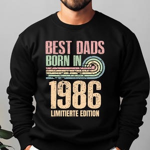 Op de afbeelding: Een zwart sweatshirt met lange mouwen en een grafische print met de tekst BEST DADS BORN IN 1986 LIMITIERTE EDITION. Het ontwerp bevat een verweerde, meerkleurige zonsondergangboog in roze, groen en blauw boven het grote jaartal 1986.